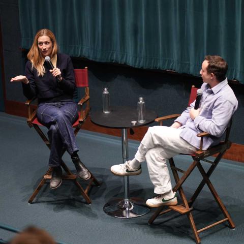 Two people seated in directors chairs on a stage holding microphones having a conversation.