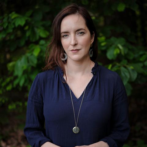 Rebecca Makkai wearing a navy blue shirt, gold pendant, and hoop earrings with green tree in the background. 