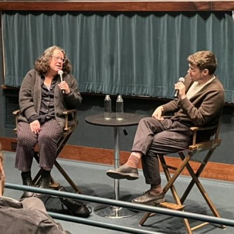 Two people sitting on a stage while sitting in wood frame, director-style chairs, speaking to each other into microphones.