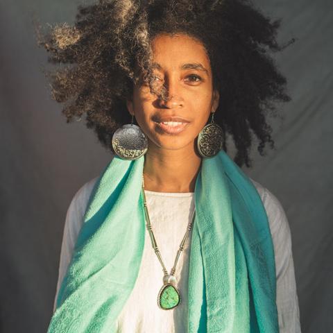 Person wearing a white long sleeve shirt, long teal scarf, necklace with large teal stone, large round gold earrings with long dark curly hair. 