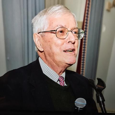 A person with grey hair and glasses wearing a sweater and tie looking to the side and speaking into a microphone.