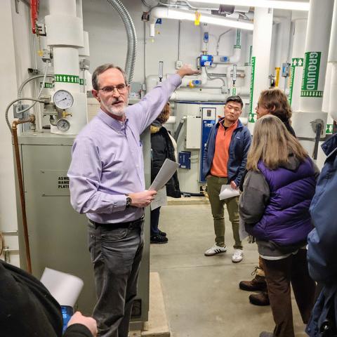 A person in a light blue button-up collared shirt lecturing to an audience while standing in a well lit mechanical room with pipes and systems in the background.