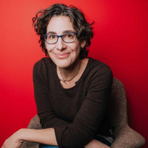 A portrait of Sarah Koenig, a person with medium-long brown hair, a dark sweater, and glasses. Koenig is sitting against a red background.