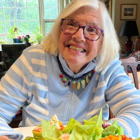 A photo of Marian Heath Mundy, a person with long gray hair, glasses, and a blue shirt. Mundy is sitting at a table with flowers on it.