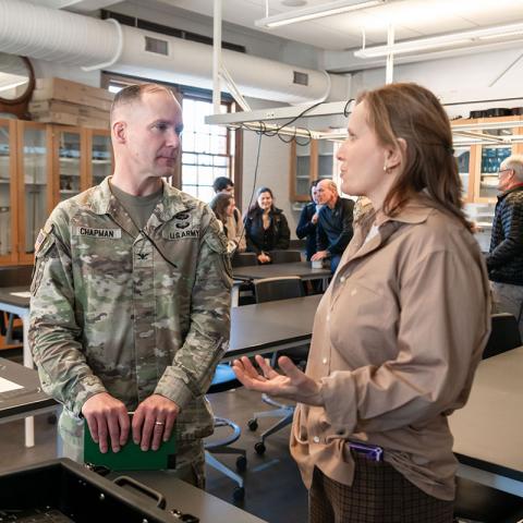 Two people standing in a classroom speaking to each other with other people and classroom tables in the background. One of the people speaking is wearing a camouflage, military uniform. 