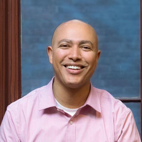 Person smiling and sitting on a stool in front of a dark-brown framed window wearing a button-down, light pink, collared shirt.