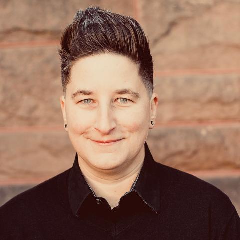 A portrait of a person in front of a red brick wall with tall brown hair and a black shirt.