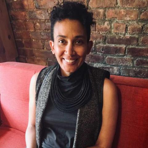 A person with short black hair sits on a red chair smiling at the camera.