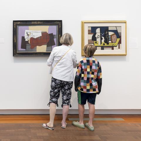 Adult woman and child stand with their backs to the viewer, looking at modern paintings in an art gallery.