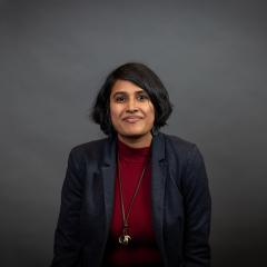 A person with medium length dark hair, dark skin, smiling, wearing a dark blue blazer, red mock-neck shirt, and long necklace with a gold pendant.