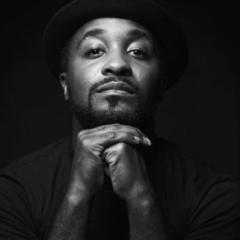 A portrait of Darrell Grand Moultrie, a person wearing dark clothes and a dark fedora. Their hands are folded and they are looking at the camera.