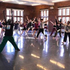 A person teaching a dance class in a large brick studio filled with other people.