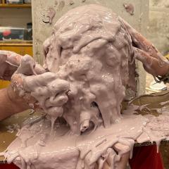 A table with a sculpture head being slathered in wet clay by messy hands.